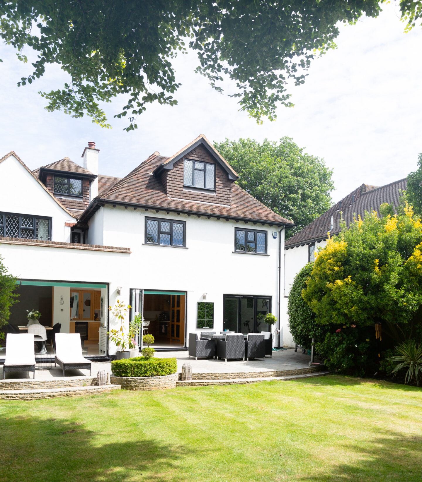 Family home featuring bi-folding doors to a paved patio and lawned garden with garden furniture. 