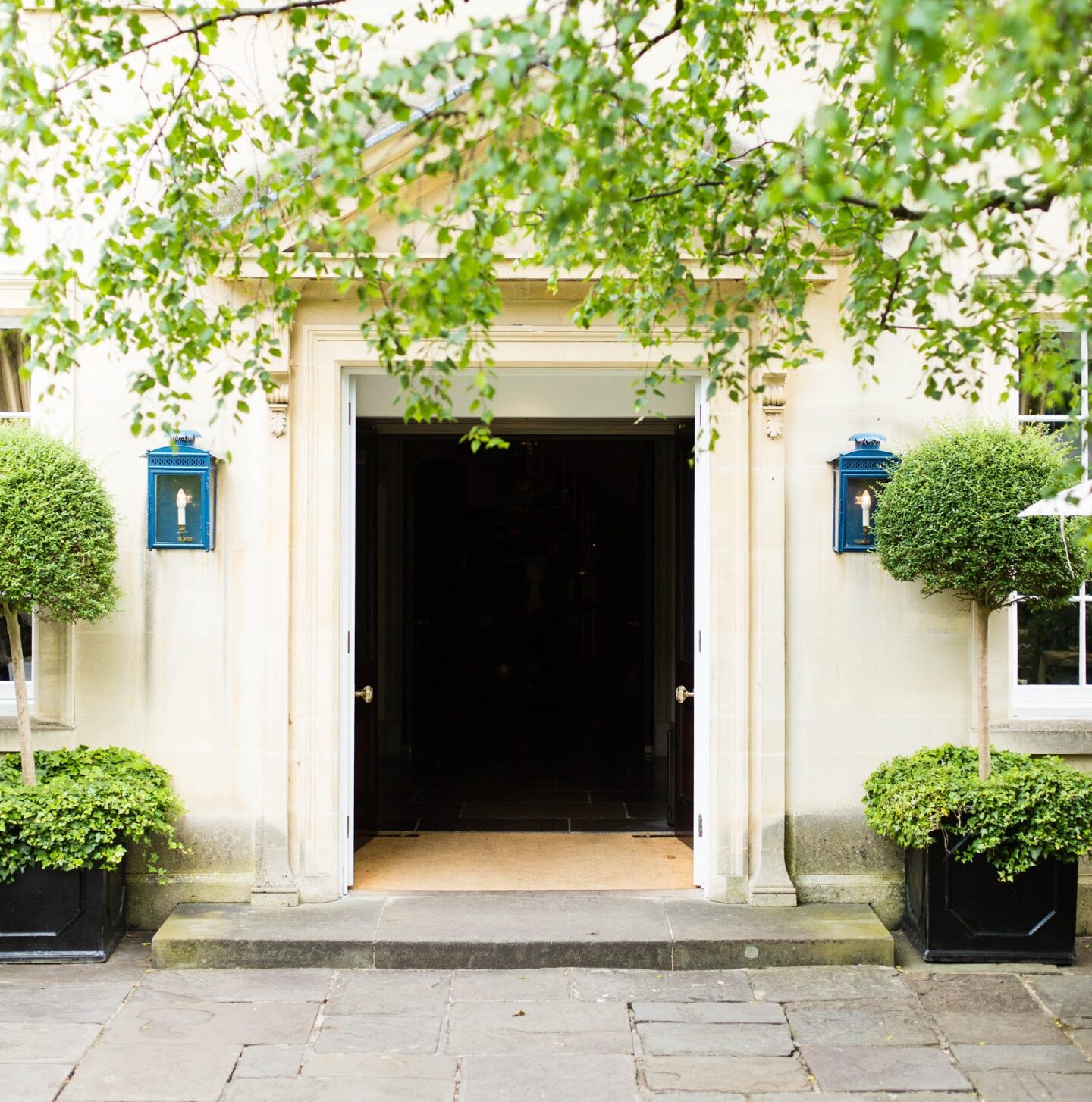 Large hall entrance with tree foliage and blue exterior lantern-style wall lights on both sides of the main door.