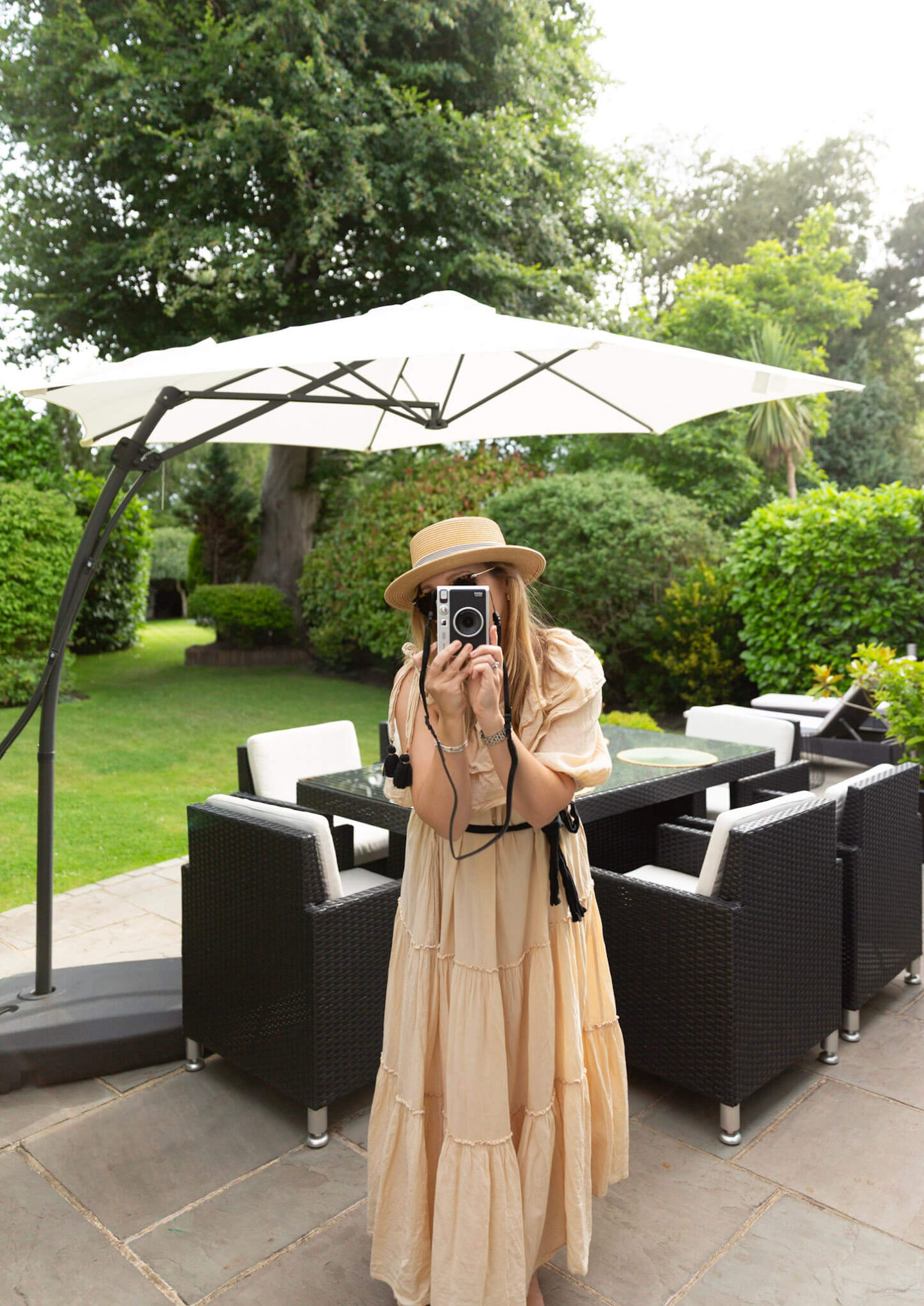 A woman holding a Fujifilm Instax Mini Evo Instant Hybrid Camera in a leafy garden with an outdoor table in the background