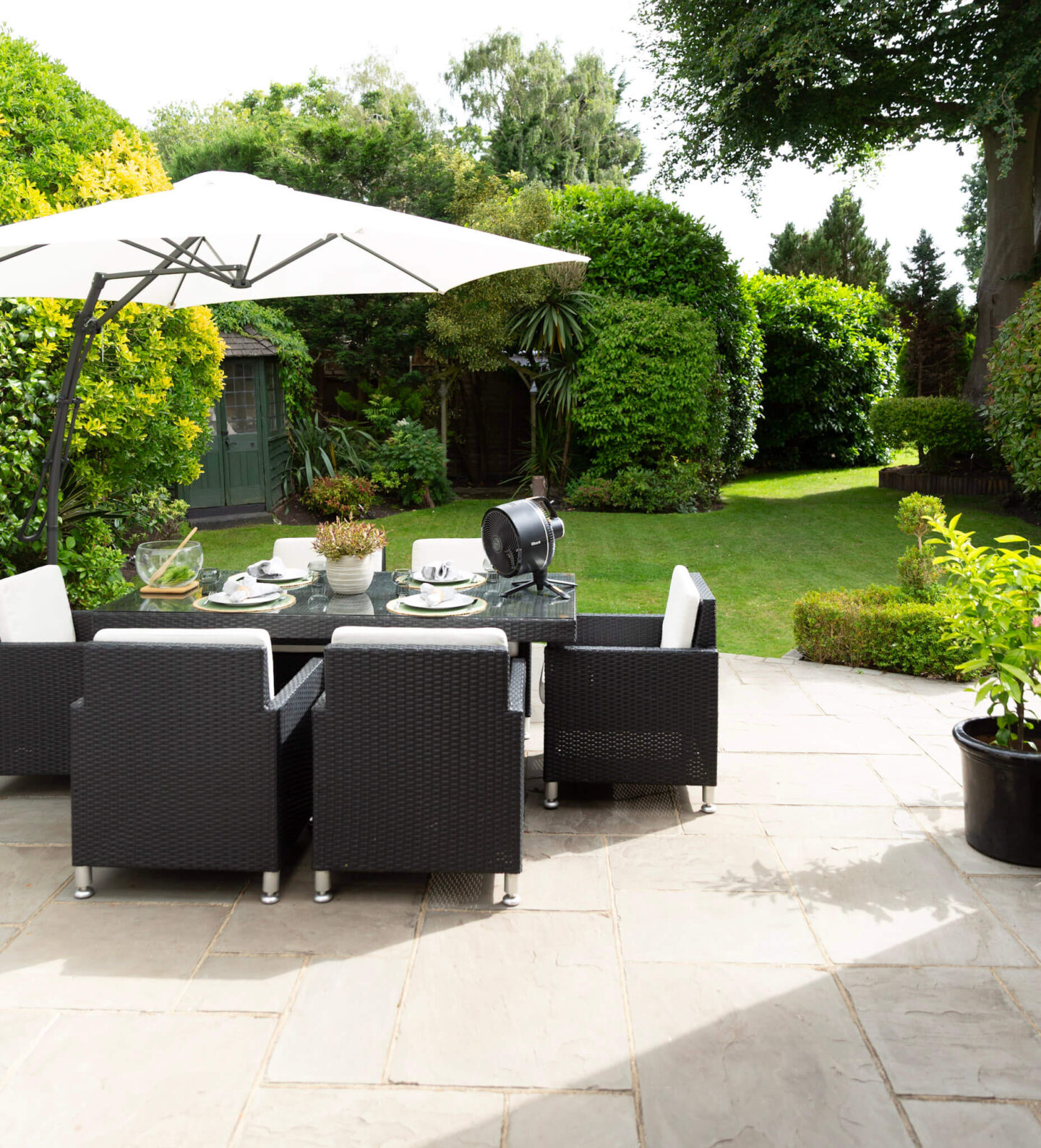 Summer garden dining setup with a portable table fan, outdoor tableware and a lush garden in the background